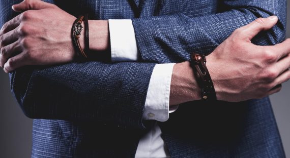 Fashion portrait of young businessman handsome model man dressed in elegant blue suit with accessories on hands posing on gray background in studio.  Keeping crossed hands